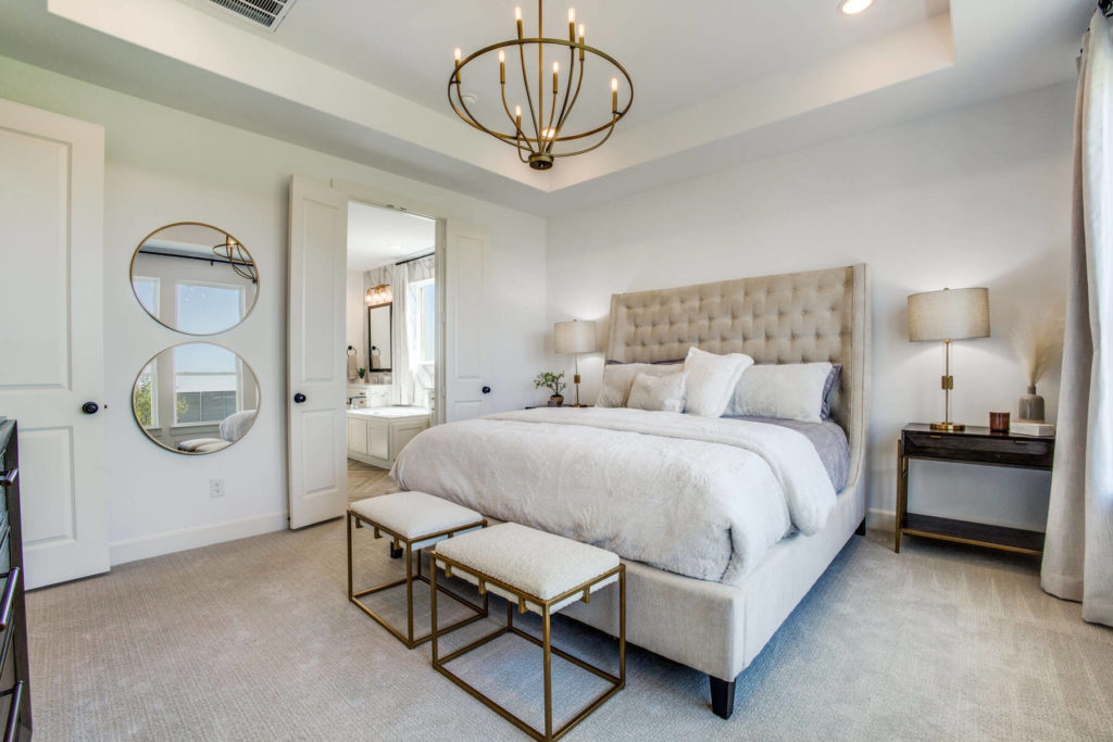 A bedroom in a newly built home in McKinney, complete with a comfortable bed, dresser, mirror and an elegant chandelier.
