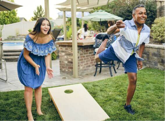 A man and woman playing cornhole in a backyard surrounded by nature.