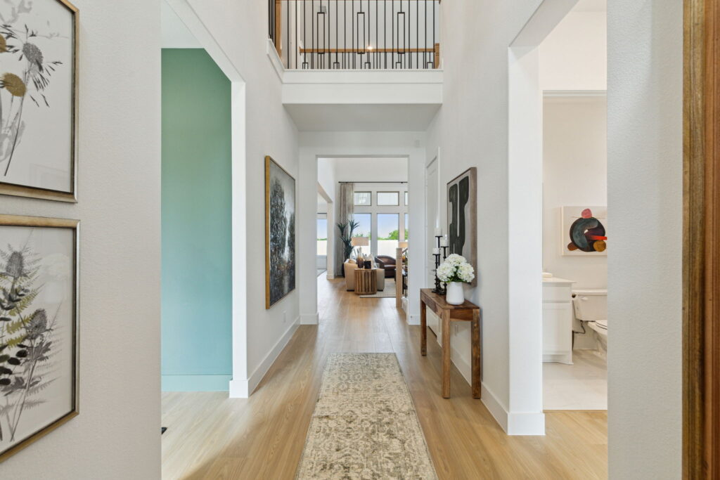 A bright, modern McKinney home corridor with art on the walls, a beige runner on the wooden floor, leading to a living area with visible upper-level balcony.