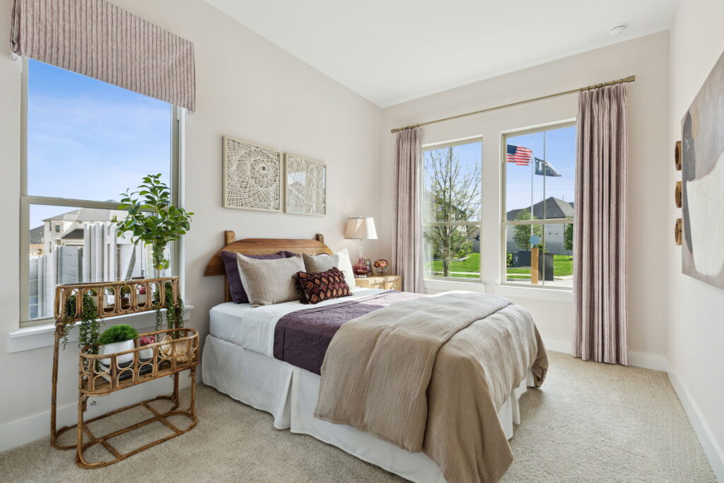 A neatly arranged bedroom in a new home with a large window displaying an outdoor view with an American flag, featuring a beige and purple bedding set, decorative pillows, and a small plant rack.