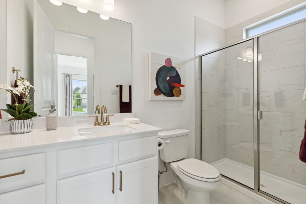 Modern bathroom in a new home featuring a white vanity, sink with gold fixtures, glass shower, and a decorative painting on the wall in McKinney, TX.