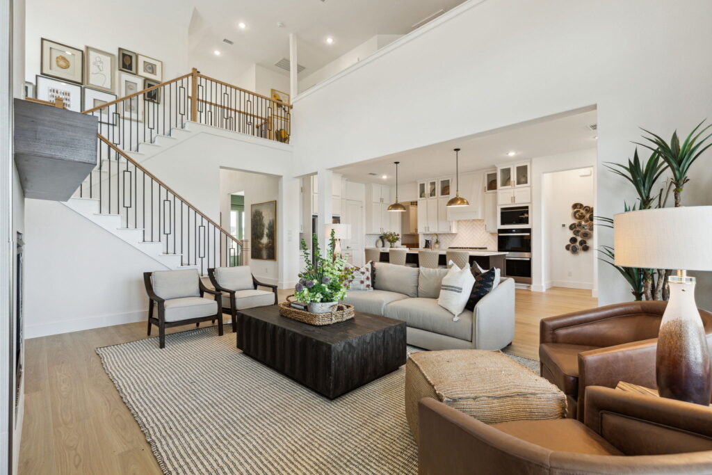 Modern open-plan living room in a New home featuring a staircase, sleek kitchen in the background, and elegant seating area with beige and brown furnishings.