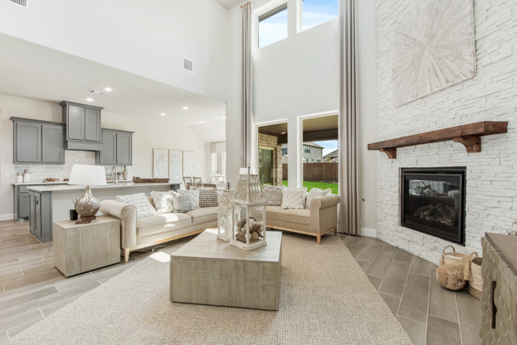 Spacious living room with high ceilings, a white stone fireplace, and adjoining kitchen by Bloomfield Homes, featuring large windows and neutral tones.