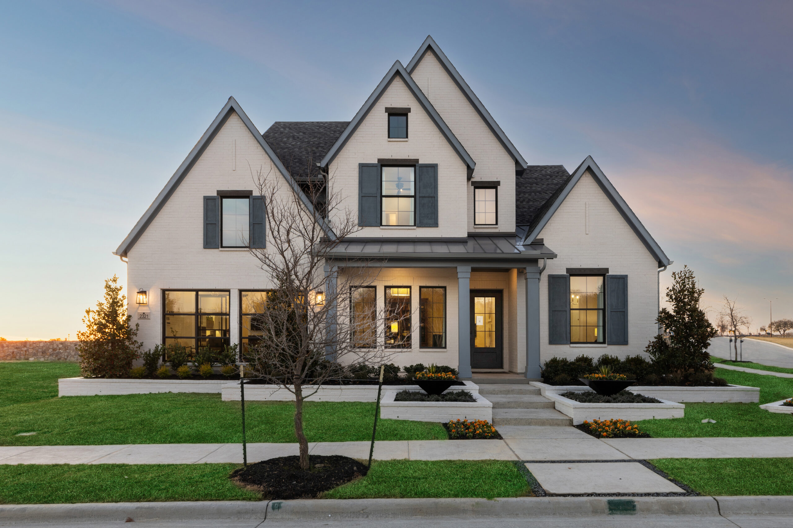 White two-story modern house with gray shutters, large windows, a gabled roof, front porch, manicured lawn, and small landscaped garden beds at sunset—discover similar Painted Tree homes for sale in the sought-after Painted Tree McKinney community.
