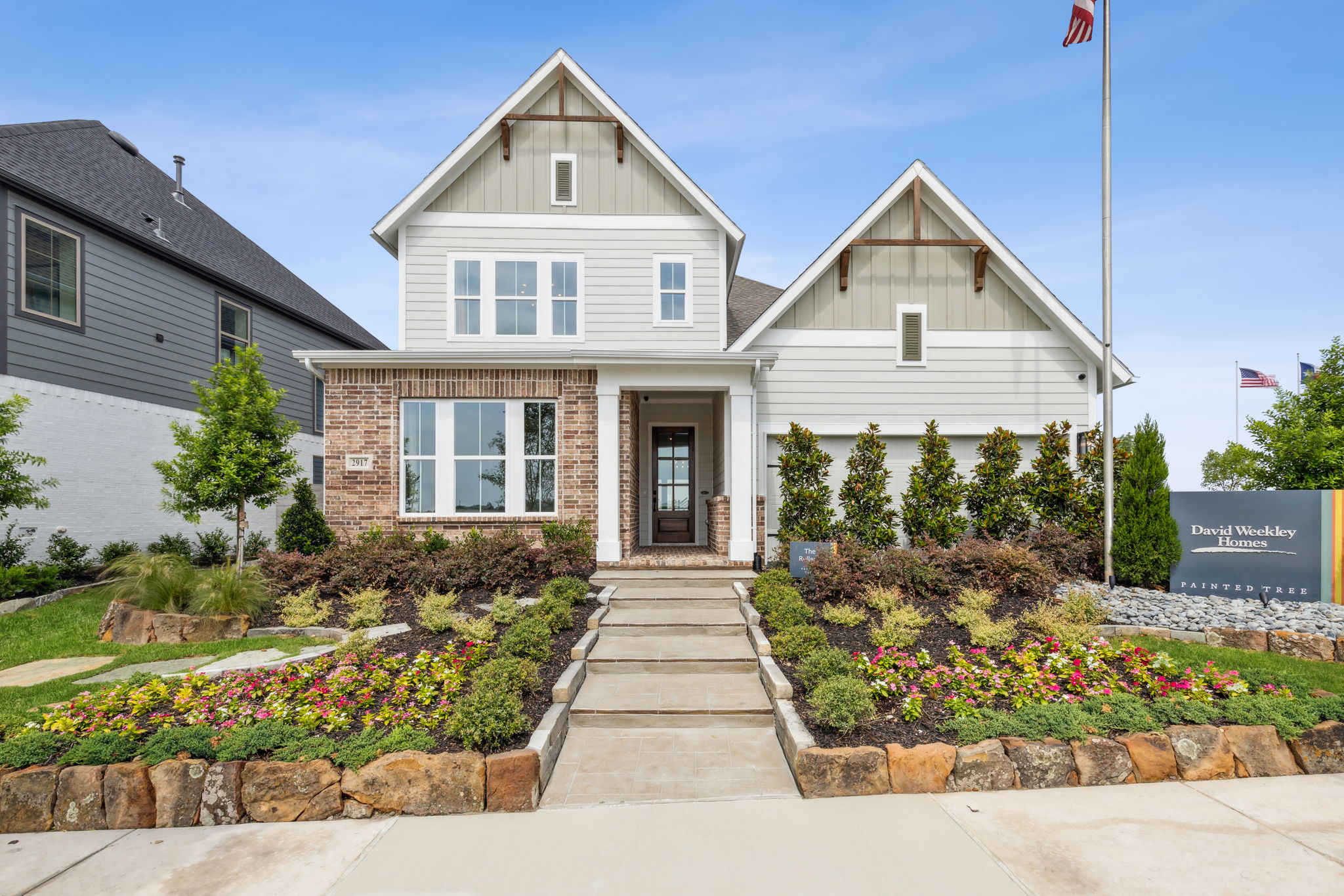 Modern two-story house with white siding, brick accents, and a landscaped front yard in Painted Tree McKinney, featuring flower beds, shrubs, and a stone walkway leading to the front door. Discover stunning homes for sale in McKinney Texas.
