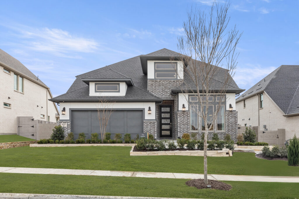 Discover your new home in this modern two-story house featuring dark brick and light siding, a sloped roof, and a spacious two-car garage. Located in the serene community of Painted Tree, TX, it boasts a front yard with trimmed grass, small shrubs, and a bare tree.