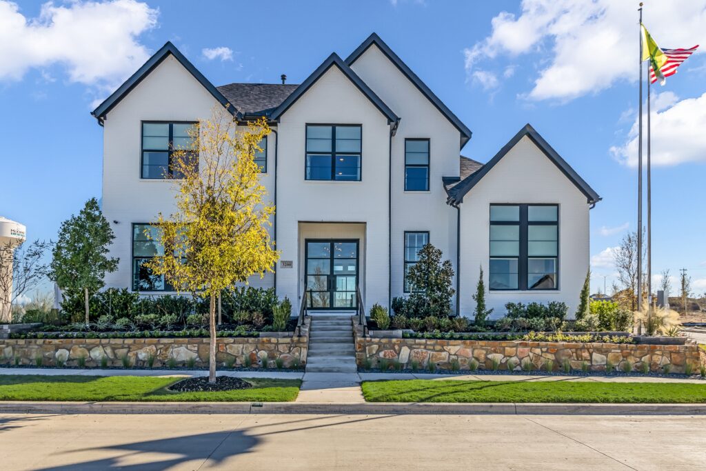 This modern two-story house in McKinney, TX, features a stone foundation, large windows, and a beautifully landscaped front yard. A proud American flag waves on the right. It's the perfect new home for sale.