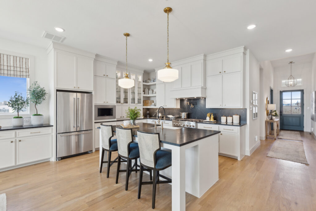 Discover a modern kitchen in one of the new McKinney homes by Southgate Homes, featuring white cabinets, a central island with stools, pendant lights, sleek stainless steel appliances, and elegant hardwood floors. Large windows fill the space with abundant natural light.