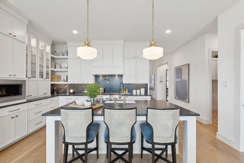 Modern kitchen in new Southgate Homes build, featuring white cabinets, a black countertop island with three cushioned chairs, pendant lights above, and wooden flooring. Visible appliances and decor on shelves complete this chic McKinney residence.