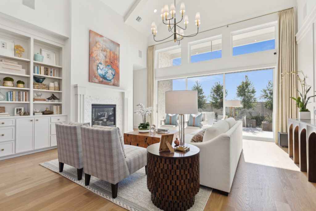 A contemporary living room in McKinney, TX, features a white sofa, plaid armchairs, and a wooden coffee table. A fireplace and built-in shelves grace the left side. Large windows reveal a patio and lush trees outside, perfect for new homes seeking charm and comfort.