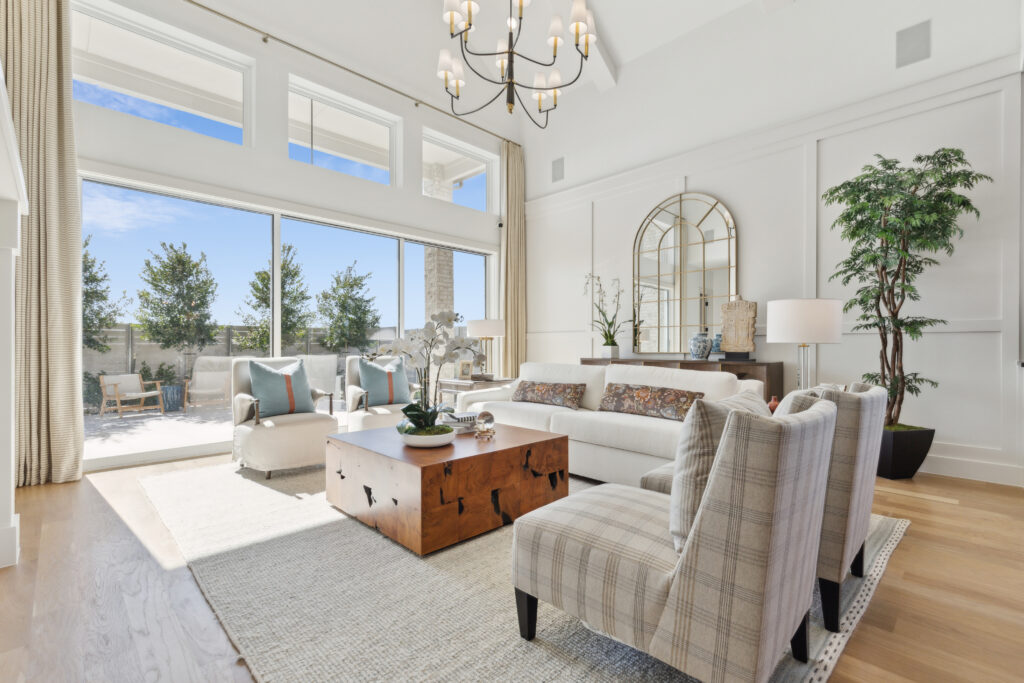 In McKinney, TX, this bright living room by Southgate Homes boasts a high ceiling with a chandelier, large windows, and glass doors. Features include white sofas, plaid chairs, a wooden coffee table, and an expansive mirror that enhances the outdoor view.