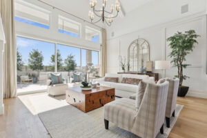 In McKinney, TX, this bright living room by Southgate Homes boasts a high ceiling with a chandelier, large windows, and glass doors. Features include white sofas, plaid chairs, a wooden coffee table, and an expansive mirror that enhances the outdoor view.