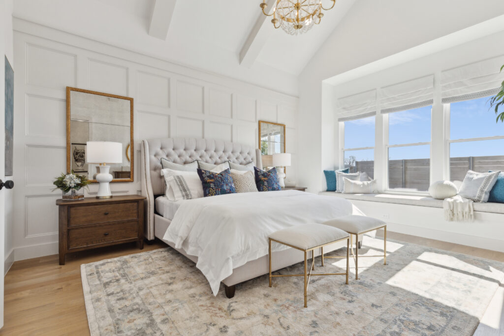 Bright bedroom in a McKinney, TX home, featuring a tufted bed, patterned rug, and bench. Two bedside tables with lamps, large mirrors, and a window seat with cushions make it cozy. A chandelier hangs overhead in this inviting new space.