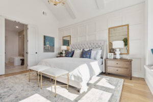 Discover a bright bedroom in a Southgate Homes new residence in TX, featuring a large bed with a tufted headboard, two benches, elegant nightstands with lamps, and artwork adorning the walls. An open door seamlessly connects to the inviting bathroom beyond.