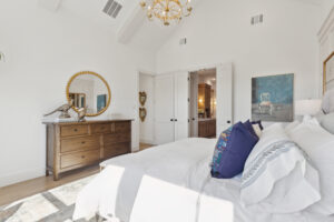 Bright bedroom in a TX new home by Southgate Homes, featuring white walls and bedding, a wooden dresser with a round mirror, and a chandelier. A doorway reveals part of an adjoining room with wooden cabinets.