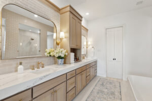 Discover the elegance of this Southgate Homes bathroom, featuring a long double vanity, marble countertop, and gold fixtures. Adorned with a floral arrangement and large mirrors with wall sconces, it boasts beige tiles and a patterned rug for a luxurious touch in your new TX home.