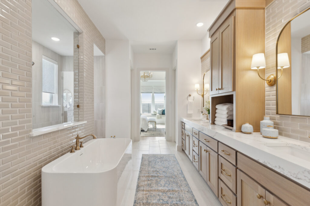 Modern bathroom in a McKinney, TX new home features a freestanding tub, wood cabinetry, dual sinks, and a marble countertop. A rug runs along the tiled floor, leading to a bedroom through the far doorway.