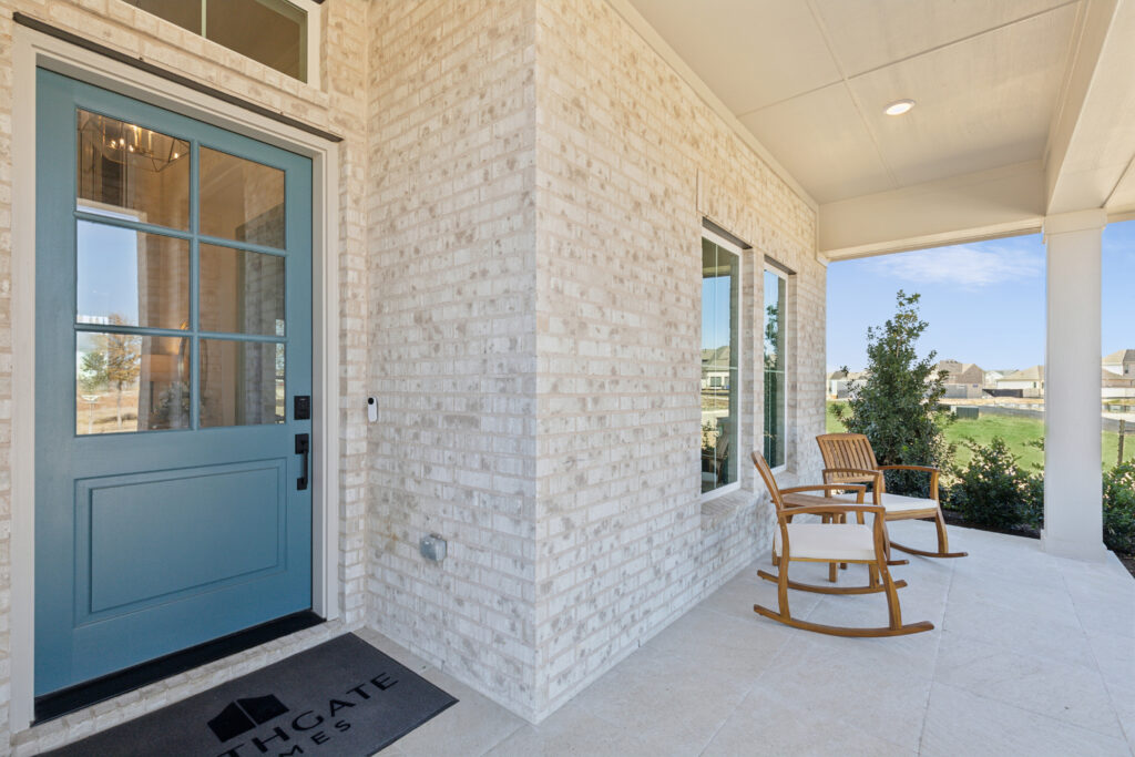 This charming front porch in McKinney, TX, features light-colored brick walls and a blue door with glass panes. Two wooden rocking chairs sit beside a welcome mat, inviting you to enjoy the lush view. A perfect touch of Southgate Homes elegance.