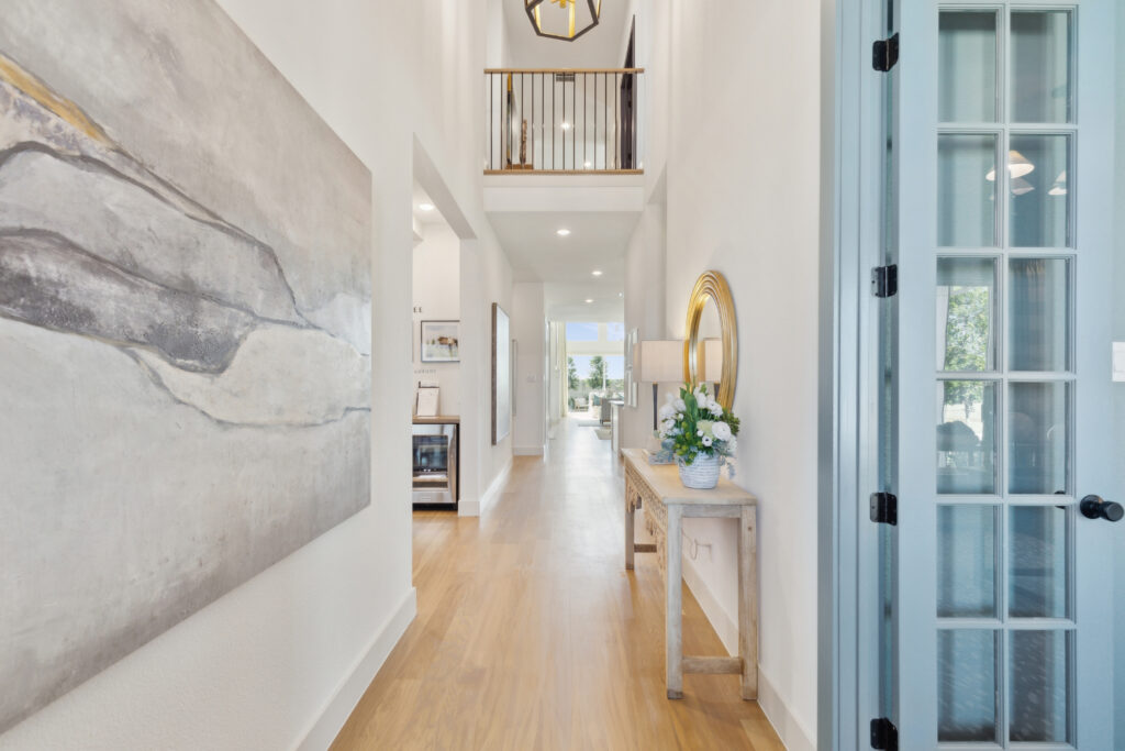 The bright hallway features a wooden floor, abstract wall art, and a console table adorned with flowers. A railing and light fixture enhance the upper section. At the end is a doorway leading to one of Southgate Homes' new homes in TX, unveiling a radiant room beyond.