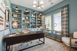 Step into a Southgate Homes-designed home office in McKinney, featuring a wooden desk, bookshelves brimming with decor, and two globe models. A patterned rug complements the large windows with checkered curtains. A chandelier and table lamp create a cozy ambiance in this inviting space.