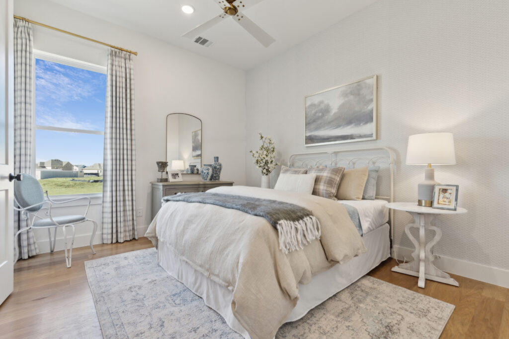 This cozy McKinney, TX bedroom by Southgate Homes features a neatly made bed, a bedside table with a lamp, and a small desk with a mirror. A chair sits by the window, offering serene views of houses. Neutral tones and decorative pillows complete the inviting decor.