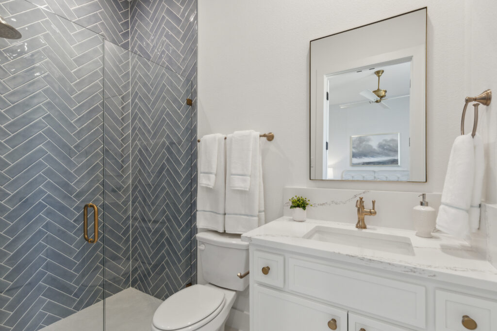 Modern bathroom in McKinney, TX, with a glass shower and blue herringbone tile. Features a white vanity with gold fixtures, a mirror, and towel racks with folded towels. Minimalist decor includes a small potted plant—a perfect addition to new homes.