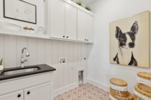 Laundry room in a Southgate Homes design features white cabinets, a black countertop sink, and woven baskets. Wall art captures the charm with a drawing of a dog over the sink and a large portrait on the right wall, embodying the style of new homes in TX.