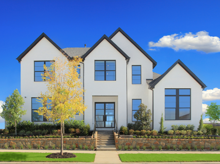 A modern two-story white house with large windows, a central walkway, landscaped shrubs, and a small tree in the front yard under a blue sky—crafted by Drees Custom Homes in the Painted Tree community of TX.