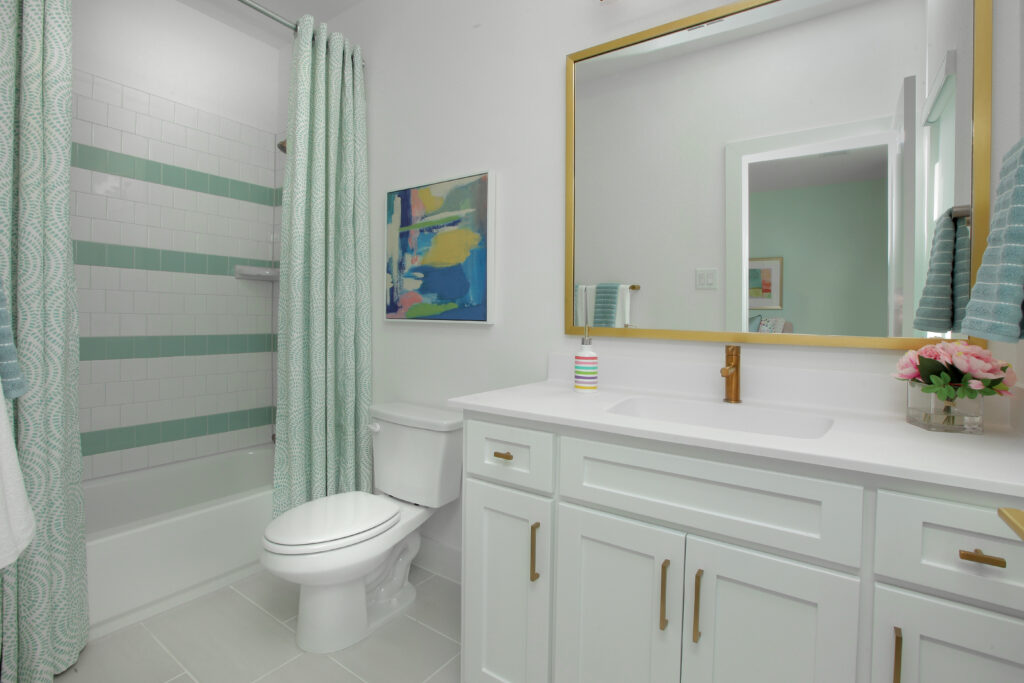 Modern bathroom in the Painted Tree community, featuring white cabinets, a gold-framed mirror, a toilet, and a shower-tub combo with green tile accents. Decorative art and flowers on the counter add charm to this Drees Custom Homes design in McKinney TX.
