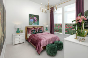 A bedroom in the Painted Tree community by Drees Custom Homes features a velvet mauve bedspread, green accent pillows, two green ottomans, white furniture, floral artwork, and large windows with purple curtains in McKinney TX.