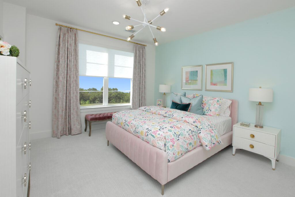 A bedroom in the Painted Tree community, featuring a pastel color scheme, a pink bed with floral bedding, two nightstands, a bench by the window, and a modern light fixture—an inviting retreat in this Drees Custom Homes new home for sale.