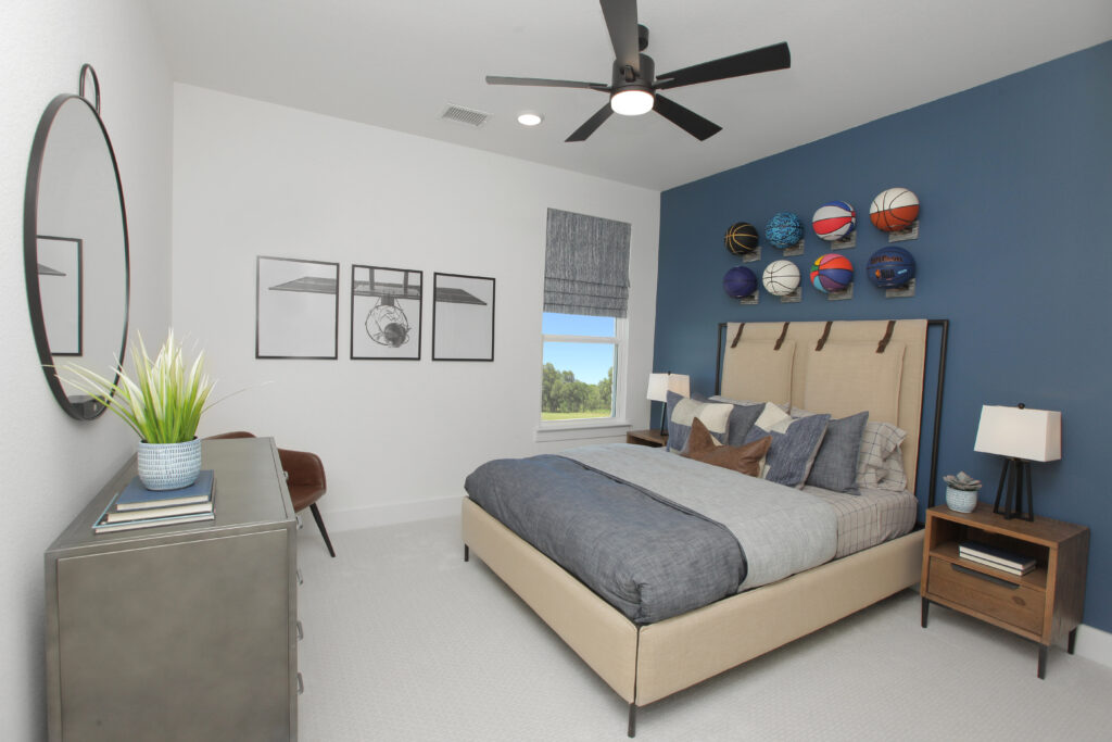Modern bedroom in the Painted Tree community, McKinney TX, featuring a blue accent wall, sports ball decor, a beige bed, dresser with round mirror, and a window overlooking trees—a perfect retreat in your new home for sale.