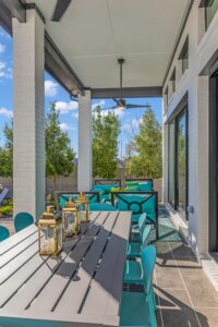 The outdoor patio by Drees Custom Homes features a long table, teal chairs, and lanterns. With large windows and lush plants in the background on a sunny day at Painted Tree, it's the perfect setting for those exploring new homes in McKinney.