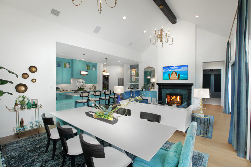 Modern open-plan living space in the Painted Tree community, McKinney TX, featuring a dining area, turquoise-accented kitchen, white sofa, fireplace, wall-mounted TV, and high ceilings with chandeliers—a stunning new home for sale.