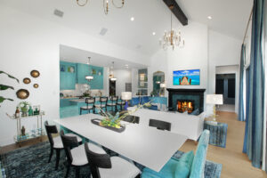 Modern open-plan living space in the Painted Tree community, McKinney TX, featuring a dining area, turquoise-accented kitchen, white sofa, fireplace, wall-mounted TV, and high ceilings with chandeliers—a stunning new home for sale.