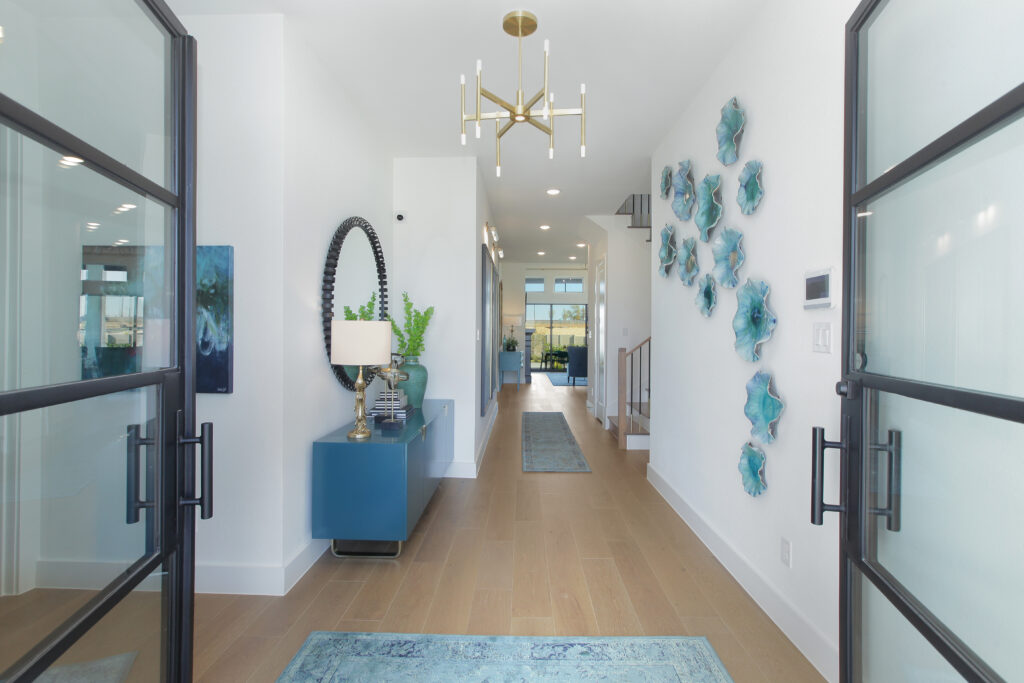 Modern entryway in a Drees Custom Homes design, featuring glass double doors, light wood flooring, blue console table with decor, wall art, and a view into a bright living space in the Painted Tree community of McKinney TX.