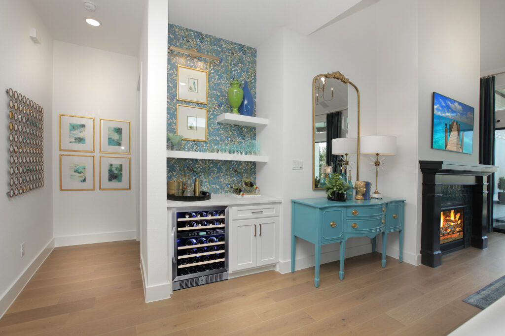 A home interior in the Painted Tree community features a small bar area, teal sideboard, mirror, lamp, fireplace, and decorative art on white walls with wooden flooring—a stunning new home for sale by Drees Custom Homes.
