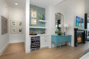 A home interior in the Painted Tree community features a small bar area, teal sideboard, mirror, lamp, fireplace, and decorative art on white walls with wooden flooring—a stunning new home for sale by Drees Custom Homes.