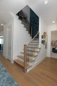 A modern staircase with light wood steps, black and gold railings, and white walls, next to a hallway with framed art in this new home for sale in the Painted Tree community, McKinney TX. A partially visible living room completes the airy space.