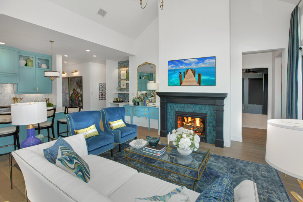 Modern living room in the Painted Tree community, McKinney TX, features blue accents, fireplace, armchairs, sofa, and a TV above the mantle with a beach scene. Dining area and Drees Custom Homes kitchen are visible in the background.