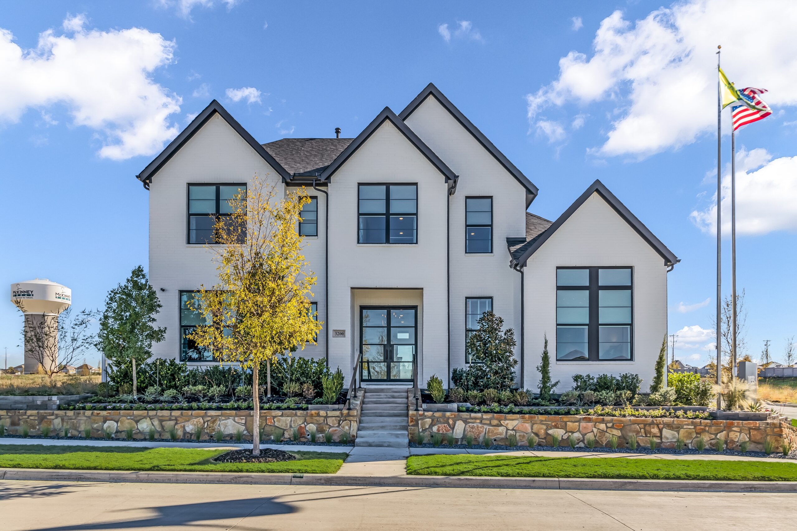 A two-story white house with a gabled roof and large windows sits gracefully on a front lawn. In the background, a painted tree and an American flag add charm, while a water tower stands tall nearby, echoing the allure of new homes in McKinney by renowned builders like Drees Custom Homes.