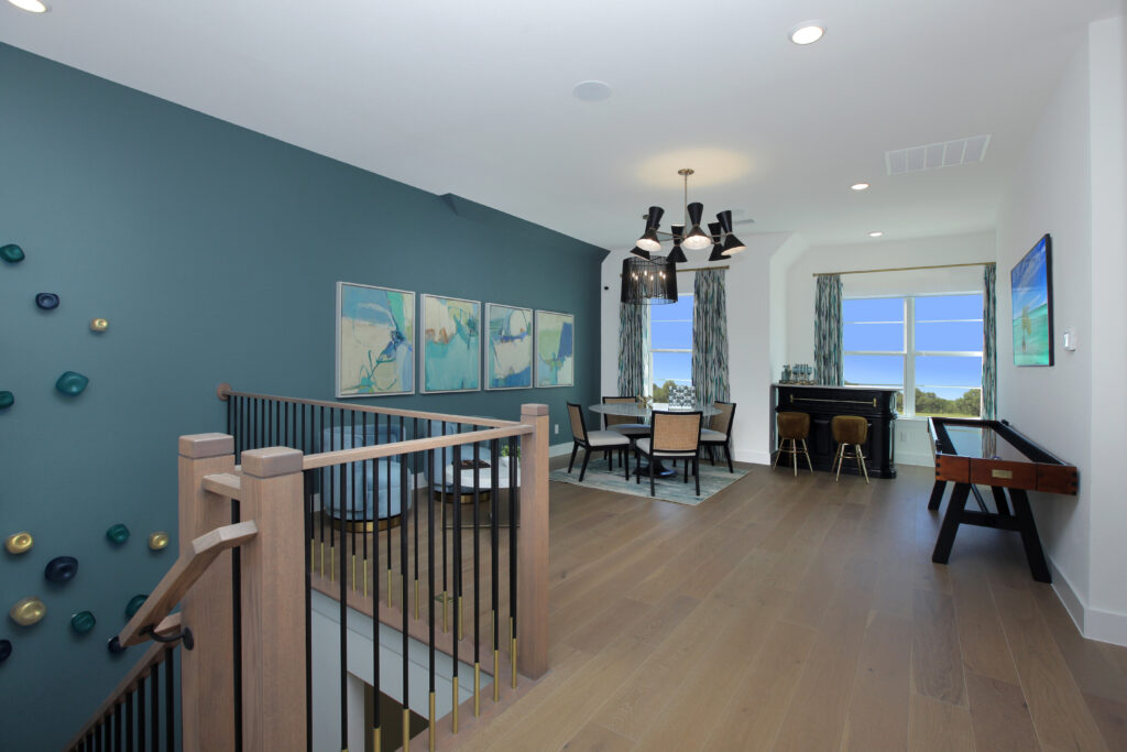 Modern upstairs game room in a Drees Custom Homes new home for sale at Painted Tree community, featuring a foosball table, dining area, wall art, teal accent wall, and large windows letting in natural light.