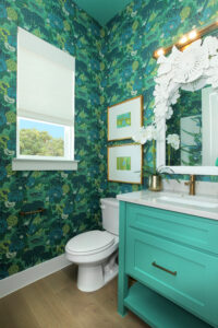 A small bathroom in the Painted Tree community by Drees Custom Homes features green floral wallpaper, a white toilet, teal vanity, decorative mirror, two framed pictures, and a window with a shade—perfect for your new home for sale.