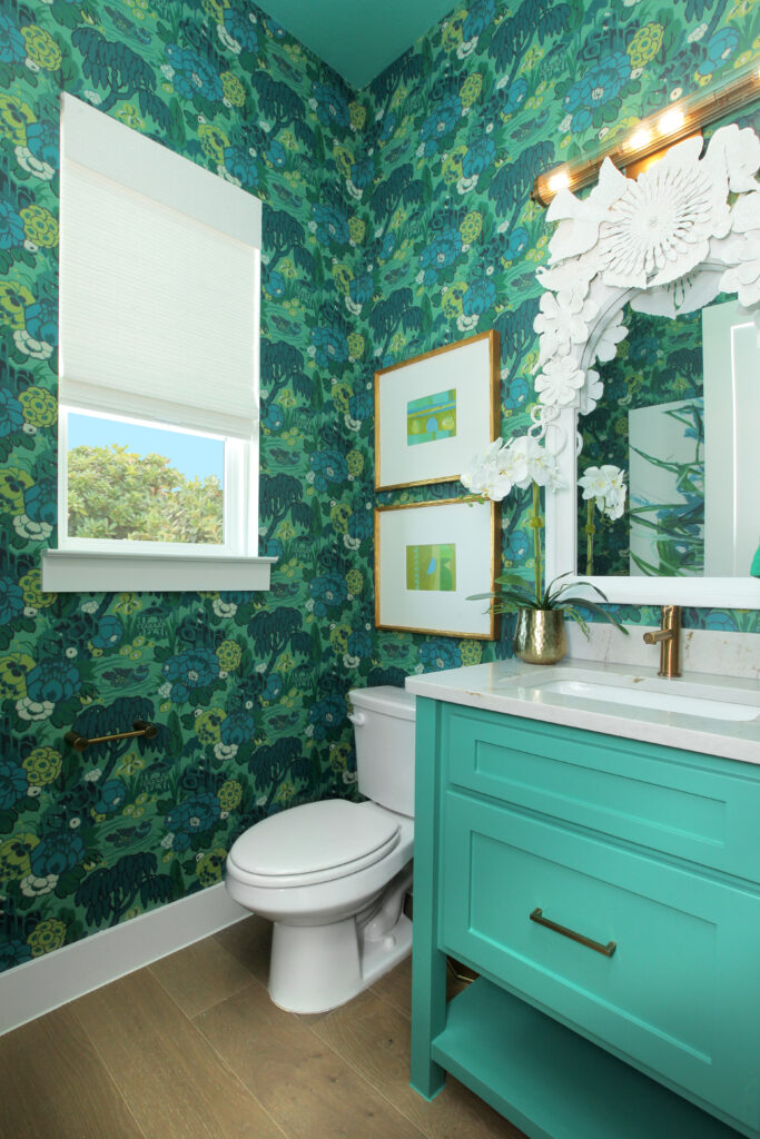 A small bathroom in the Painted Tree community by Drees Custom Homes features green floral wallpaper, a white toilet, teal vanity, decorative mirror, two framed pictures, and a window with a shade—perfect for your new home for sale.