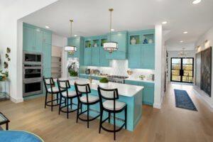 Modern kitchen with turquoise cabinets, white countertops, four barstools, pendant lights, and built-in ovens in a stunning Drees Custom Homes design; hallway and front door visible—located in the Painted Tree community of McKinney, TX.