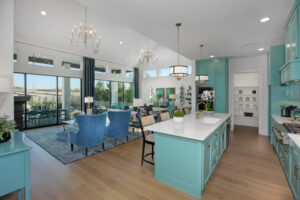 Modern open-concept kitchen and living area in McKinney, TX with turquoise cabinets, island seating, dining table, blue chairs, wood flooring, large windows, and glass doors leading to a patio—your new home for sale by Drees Custom Homes.