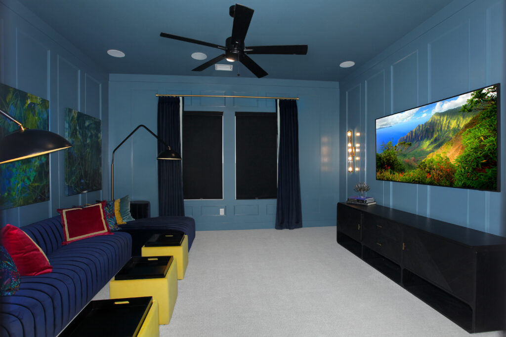 A modern home theater room in the Painted Tree community features blue walls, a large wall-mounted TV, a blue sectional sofa with colorful pillows, yellow ottomans, and dark curtains—perfect for relaxing in McKinney TX.