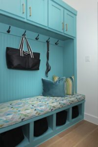 A turquoise mudroom bench with patterned cushions, throw pillows, overhead cabinets, black hooks holding a bag and leash, and storage cubbies below adds charm to this new home for sale in the Painted Tree community of McKinney TX.