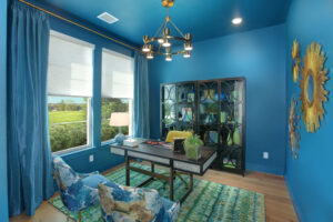 A modern home office with blue walls, blue curtains, a glass desk, patterned chairs, and sunburst wall decor—this new home for sale in the Painted Tree community, McKinney TX features large windows with a view of greenery.