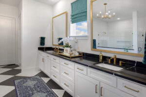 Modern bathroom in the Painted Tree community, McKinney TX, featuring a double vanity with black countertops, gold fixtures, large framed mirrors, checkered tile floor, teal window shade, and decorative accents—your new home for sale awaits!.